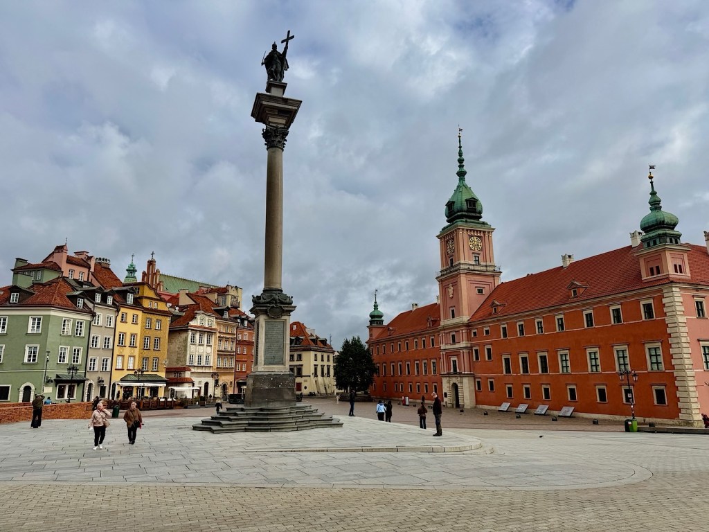A view of a square in Warsaw featuring a tall column with a statue on top, surrounded by colorful historical buildings and visitors walking along the cobblestone path.