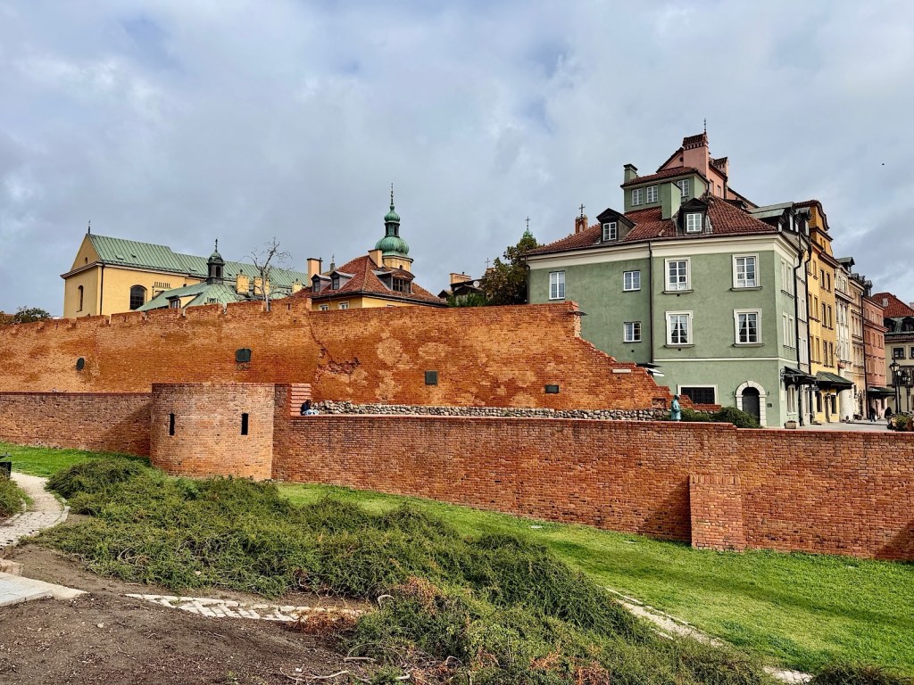 A historical brick wall with a round tower in front, surrounded by green grass and a path, alongside colorful old buildings under a cloudy sky.