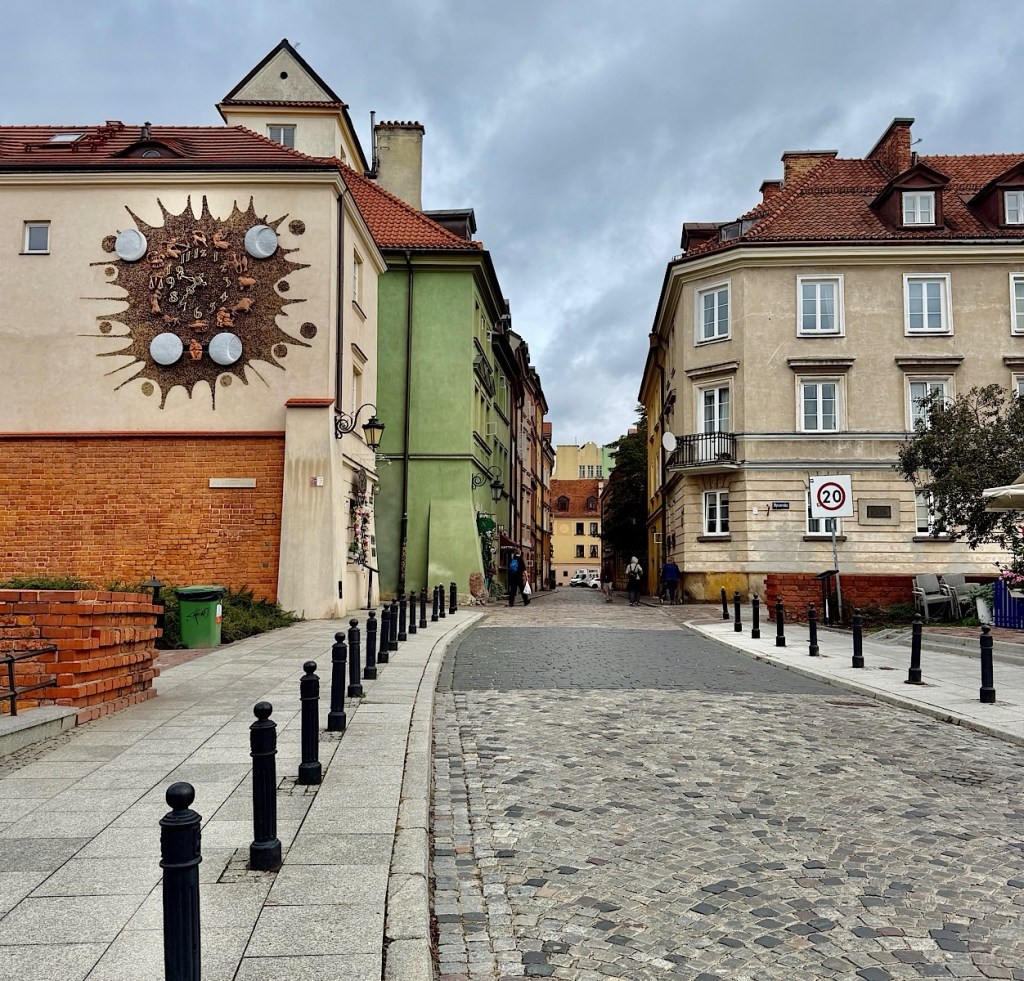 A cobblestone street with colorful buildings on either side, featuring a wall decoration of a sun-like design and several street lamps.