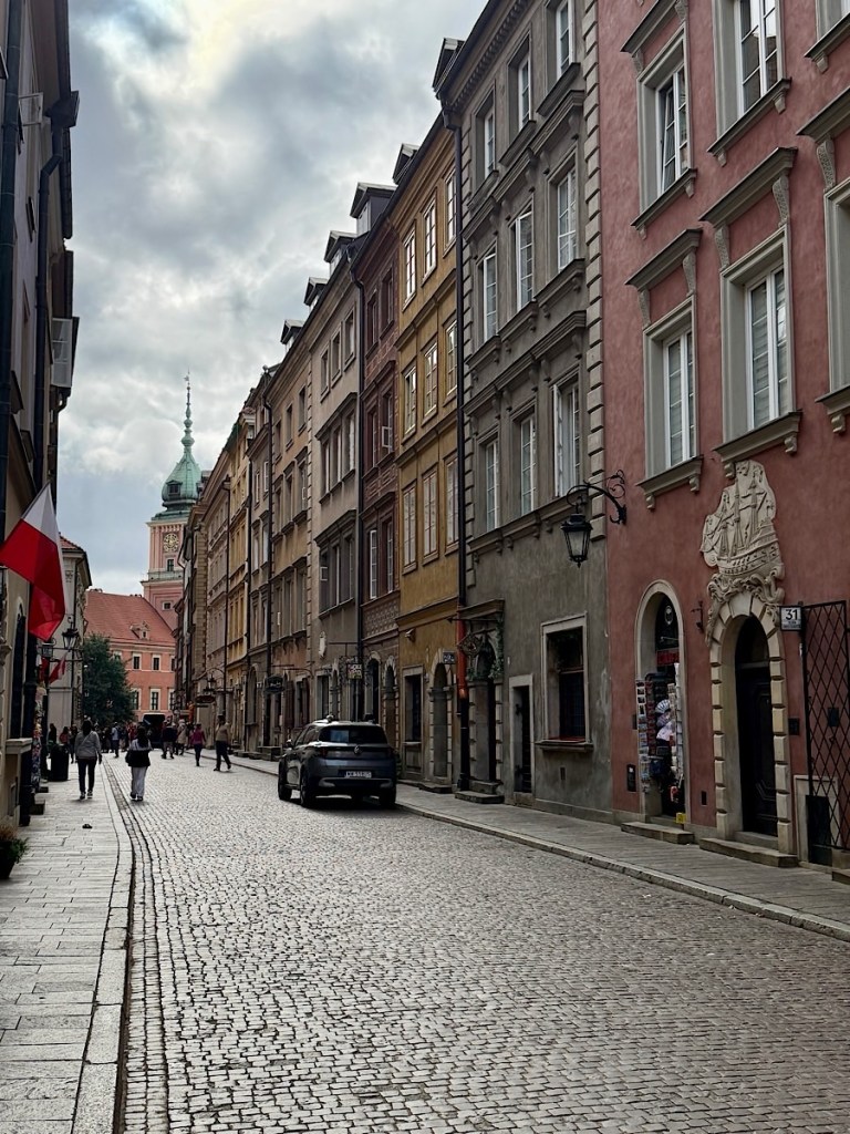 Narrow cobblestone street lined with historic buildings in warm colors, with a distant tower visible against a cloudy sky, and people walking along the street.