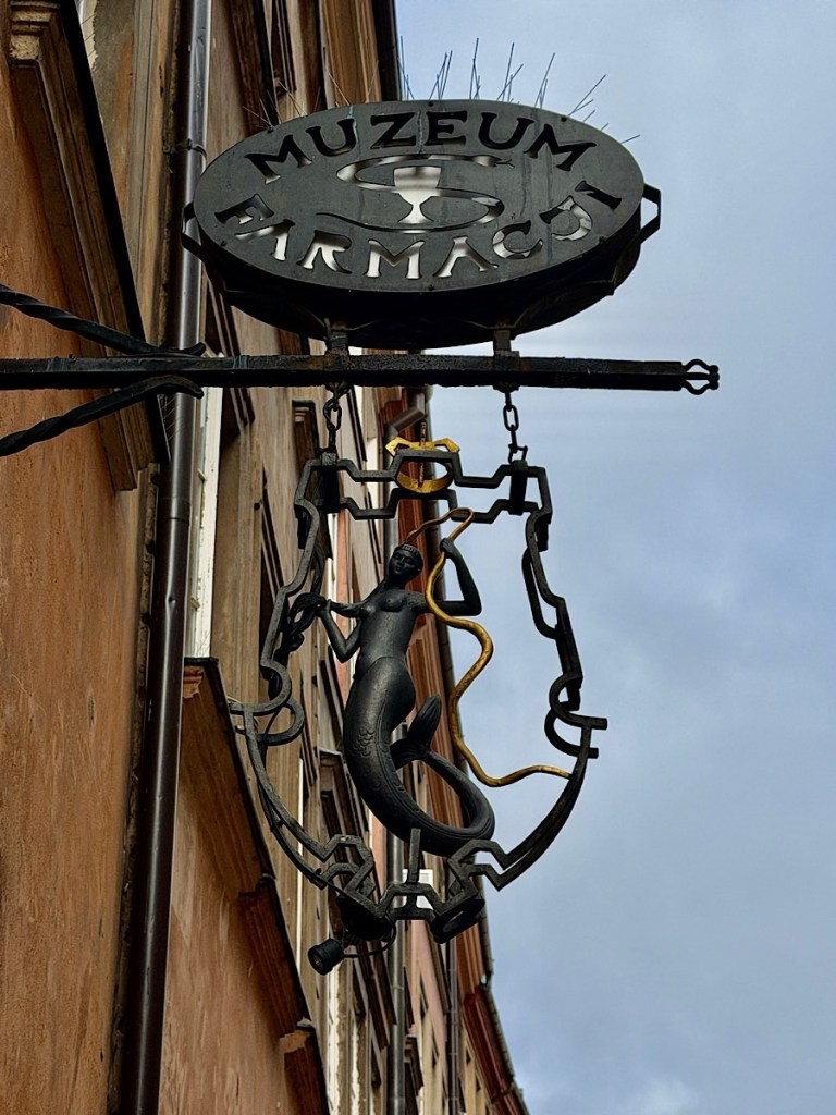A decorative sign for a museum, featuring a silhouette of a mermaid and a snakes entwined around a rod, with the word 'MUZEUM' prominently displayed above.