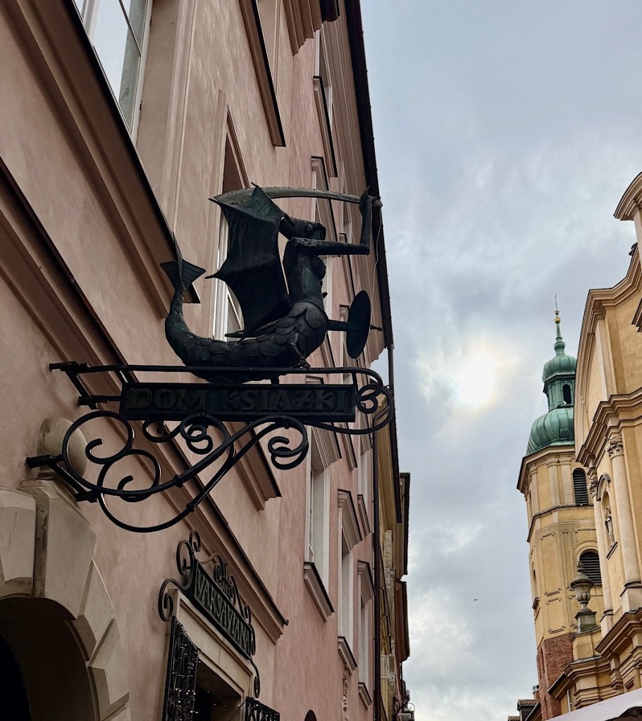 A decorative sign featuring a dragon-like creature holding a bow and arrow, mounted on a building in a narrow street with historical architecture in the background.