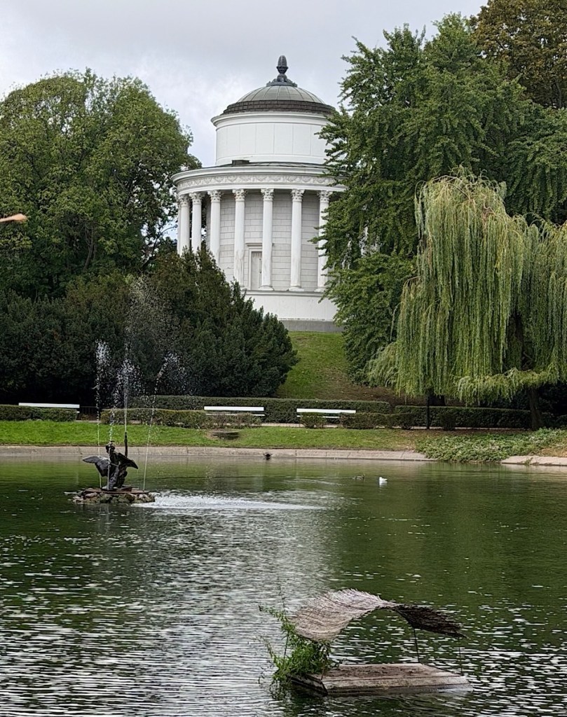 A serene park scene featuring a classical-style building on a hill, surrounded by lush greenery and a calm pond with a small fountain and a statue.
