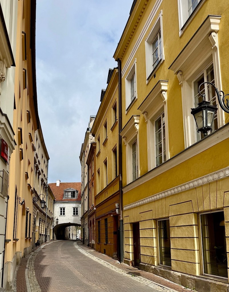 A narrow cobblestone street lined with colorful historic buildings, including a prominent yellow building, leading to an archway in the distance under a cloudy sky.
