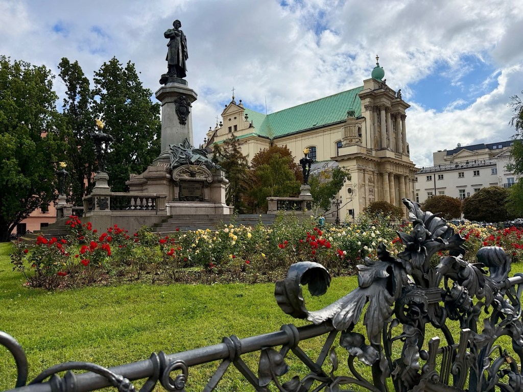 A scenic view of a park featuring a statue on a pedestal surrounded by colorful flower beds, including red and yellow roses, with a historic building and trees in the background under a cloudy sky.
