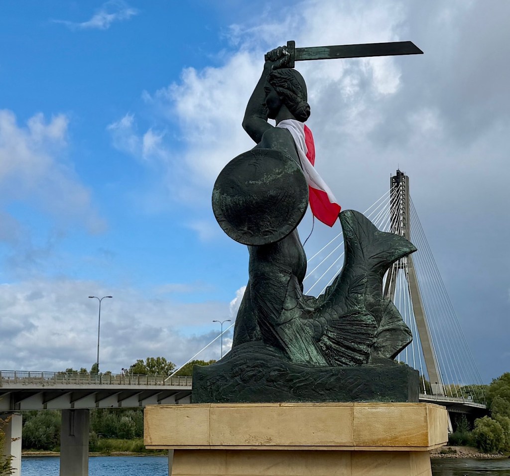 A bronze statue of a mermaid holding a sword and shield, draped in a white and red flag, with a modern bridge in the background and a partly cloudy sky.