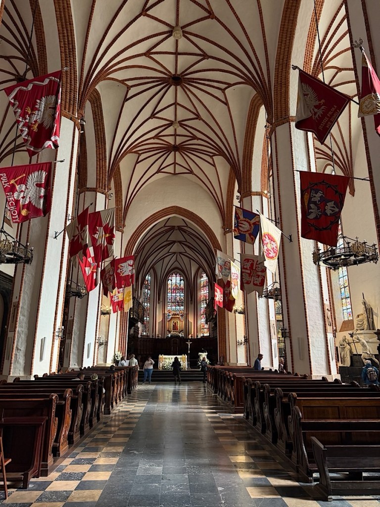Interior of a historic church featuring tall arched ceilings, wooden pews, and colorful banners hanging from the walls. The altar is visible at the end of the aisle, with stained glass windows illuminating the space.