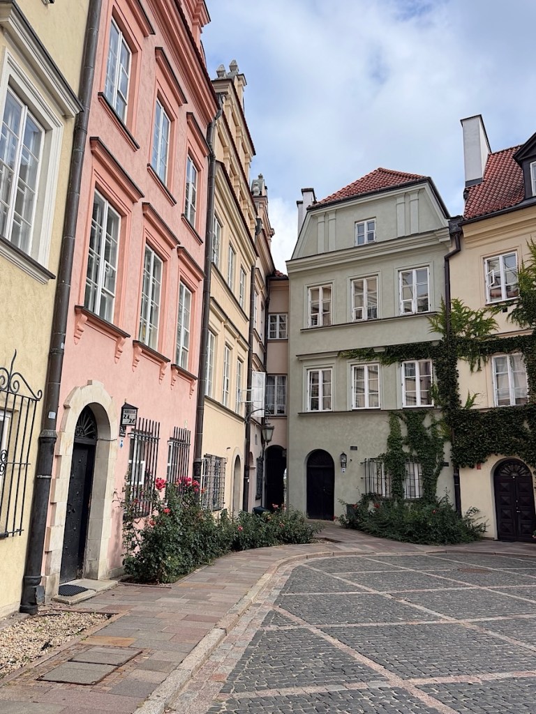 Narrow street lined with colorful historic buildings featuring pastel facades and decorative windows, with a cobblestone path and greenery in the foreground.