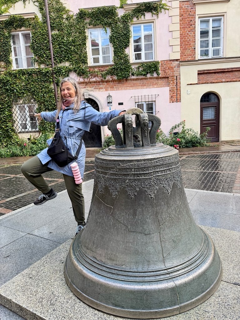 A woman joyfully poses beside a large bell sculpture adorned with decorative carvings, set against a backdrop of ivy-covered buildings.