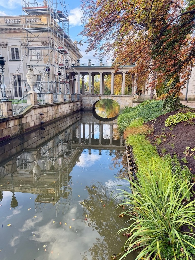 A scenic view of a canal with a bridge and classical architecture in the background. The reflection of the structure in the water is visible. Autumn foliage lines the banks of the canal.