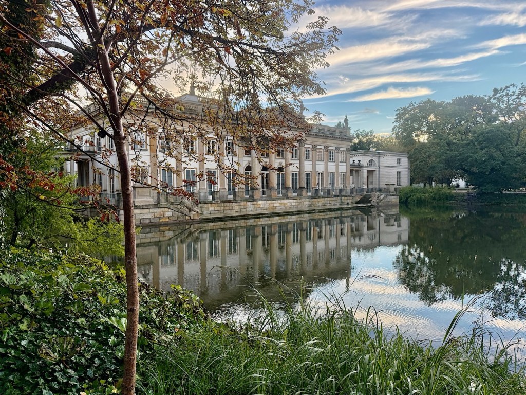 A historic building reflected in a peaceful lake, surrounded by lush greenery and autumn foliage under a cloudy sky.