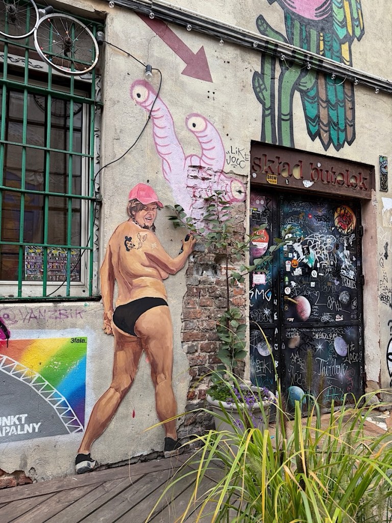 A mural depicting a person in a swimsuit with a pink cap, standing next to whimsical pink and green creatures on a brick wall, with an arrow pointing down and a graffiti-covered door beside them.
