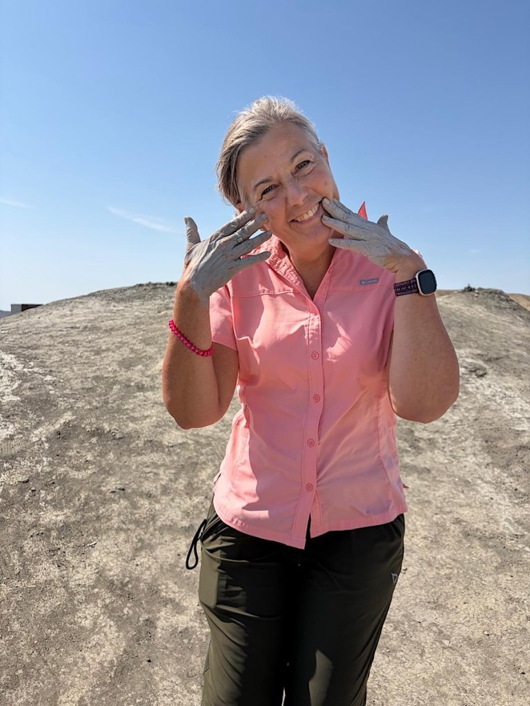 A smiling woman in a pink shirt and dark pants stands outdoors, showing her hands covered in gray mud, against a clear blue sky.
