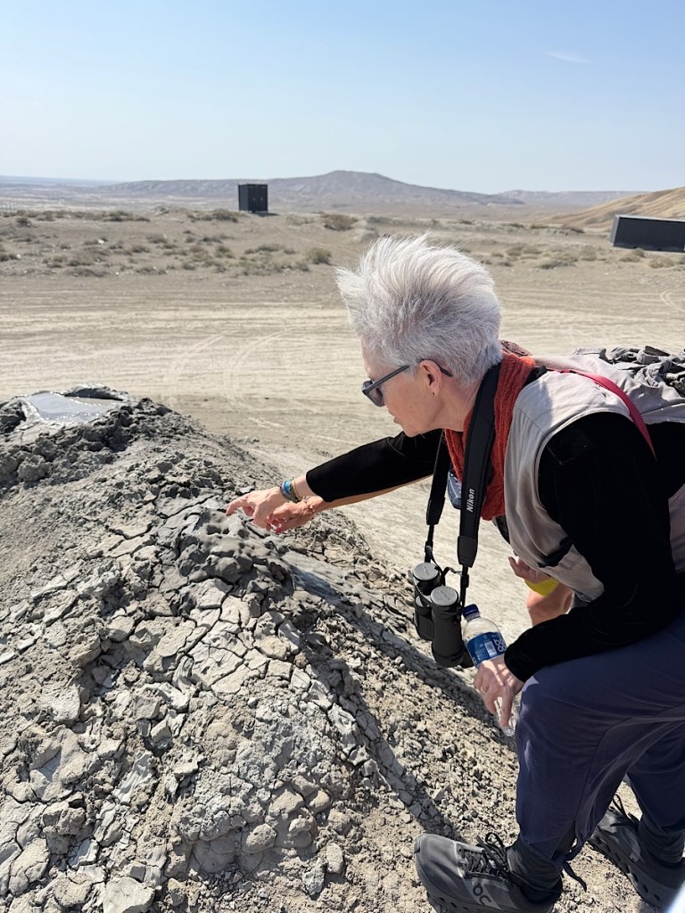 A person examining a rocky terrain with dry, cracked soil in a desert landscape, wearing a vest, binoculars around their neck, and reaching out towards the ground.