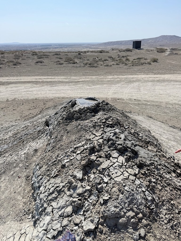 A dry, cracked earth landscape with a small mound of hardened mud and gray soil, under a clear sky. A distant black structure is visible, with sparse vegetation around.