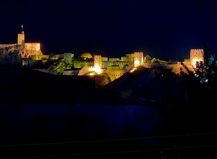 Illuminated fortress on a hill at night, showcasing towers and walls against a dark sky.
