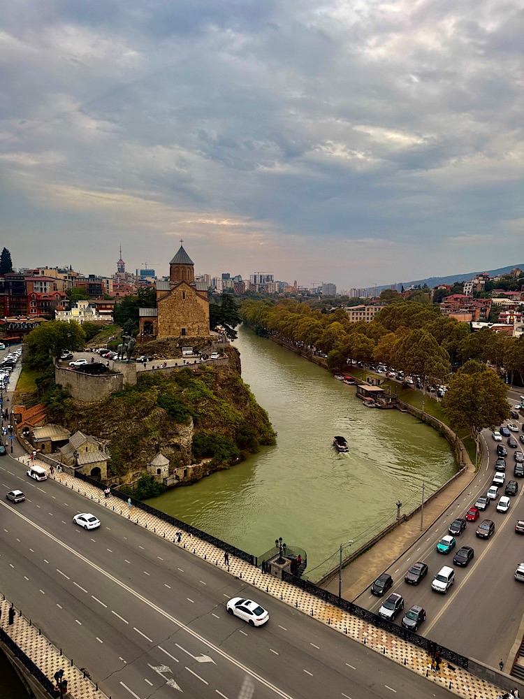 Aerial view of the Kura River winding through Tbilisi, Georgia, with a historic church on a rocky outcrop, surrounded by modern city buildings and trees along the riverbank.