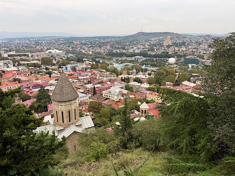 A panoramic view of Tbilisi, Georgia, showcasing colorful rooftops, lush greenery, and historical architecture, with a backdrop of hills and modern developments.