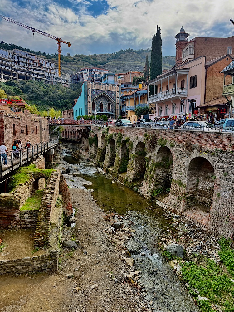 A scenic view of a riverside cityscape featuring historic buildings and arched structures alongside a gentle stream, with modern constructions visible in the background under a cloudy sky.