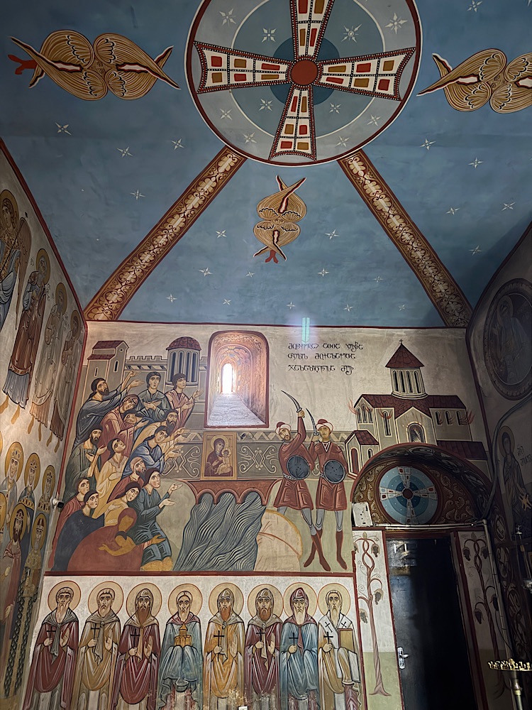 Interior of a church with intricate frescoes on the walls and ceiling, featuring religious figures, a large cross, and scenes from biblical narratives.