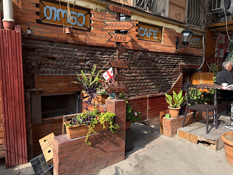 A cozy café entrance with wooden directional signs pointing to various locations, surrounded by potted plants and vibrant greenery.