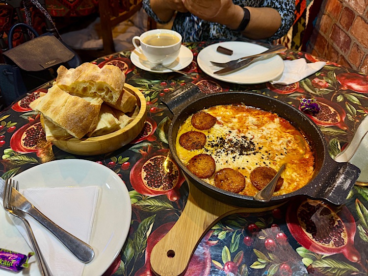 A table setting featuring a bowl of bread, a plate with cutlery, and a skillet dish with melted cheese and sausage slices served on a wooden board, alongside a cup of coffee and a plate with additional cutlery.