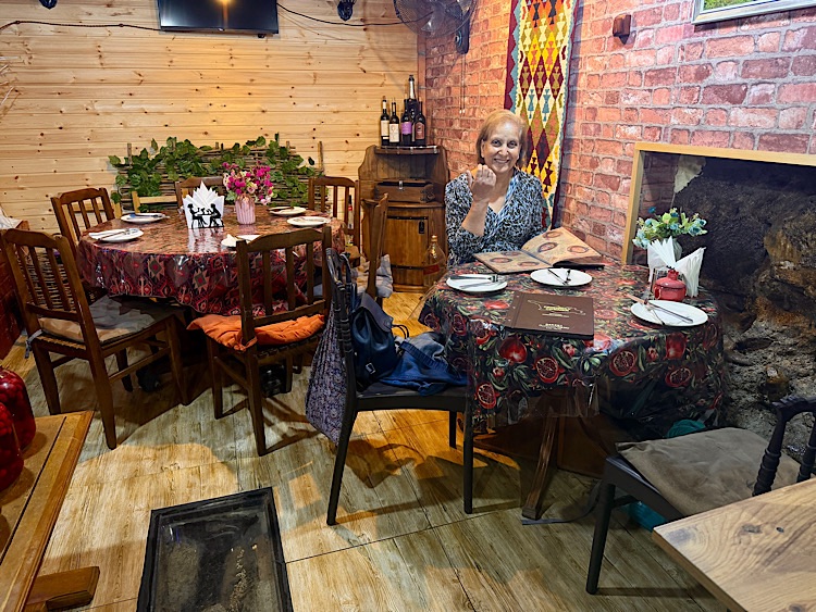 A cozy restaurant interior featuring wooden furniture, floral tablecloths, and decorative elements. A woman sits at a table with a menu, smiling while holding her finger to her lips.