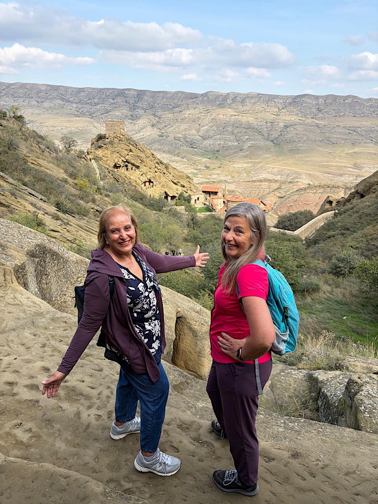 Two women standing on a rocky ledge with a mountainous landscape in the background, smiling and posing for the photo.