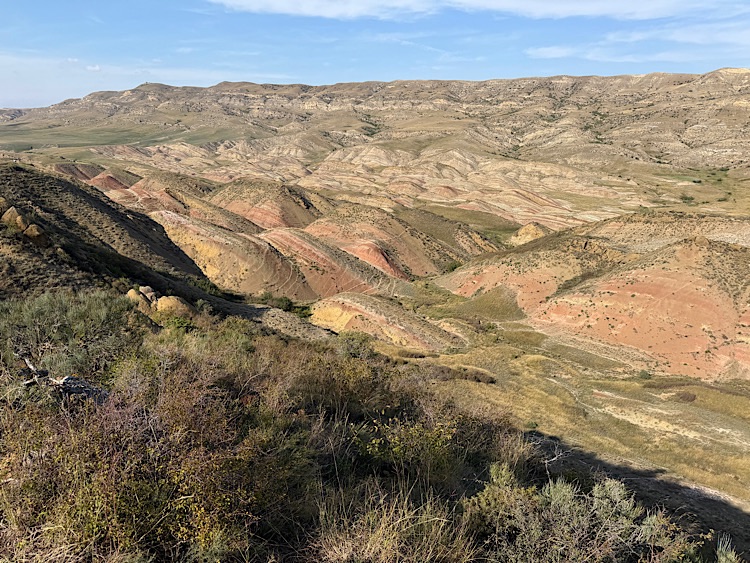 A scenic view of rolling hills and valleys featuring layered soil in various shades of brown, red, and green, under a clear blue sky.