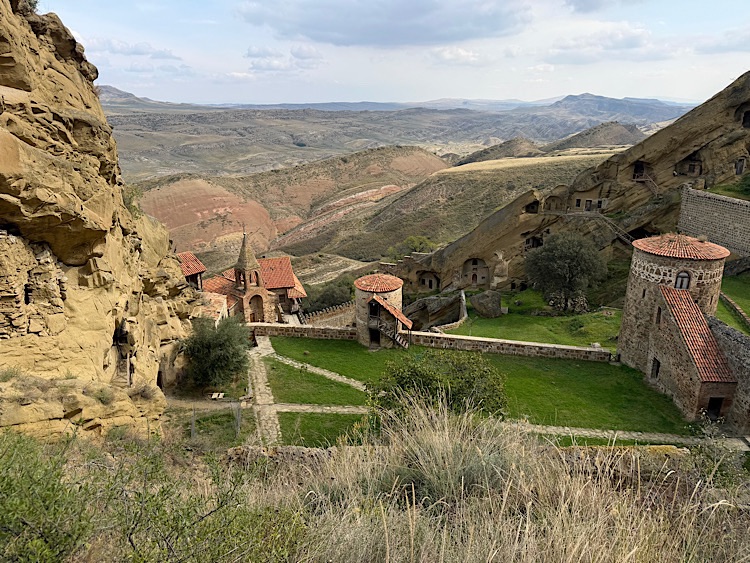 A panoramic view of an ancient monastery complex nestled in rocky terrain, with red-tiled roofs and stone walls, surrounded by rolling hills and mountains under a cloudy sky.