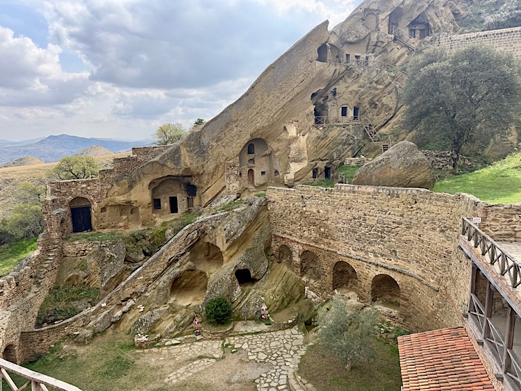 A historical rock-hewn monastery complex carved into cliffs, featuring various cave-like structures, walking paths, and greenery in the surrounding landscape.