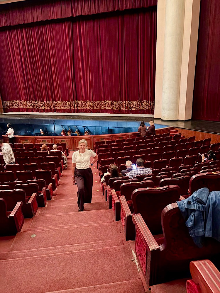 Interior of a theater with red seating and a velvet curtain, featuring a woman walking down the stairs amidst an audience.