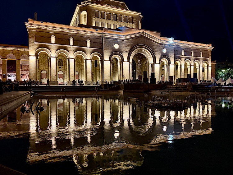 Illuminated building at night reflected in a calm body of water, with people gathered nearby.