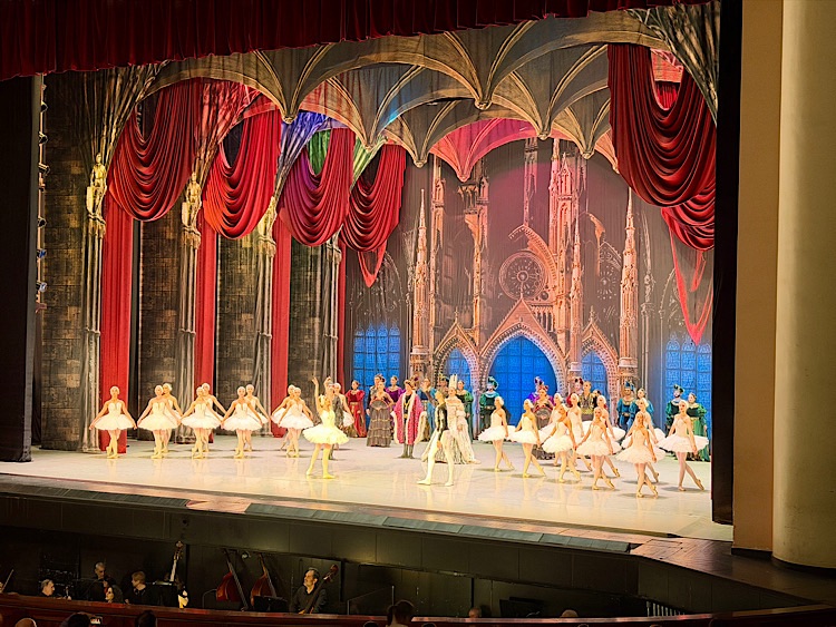 A vibrant ballet performance on stage featuring dancers in tutus, with colorful costumes and an elaborate backdrop of gothic architecture.