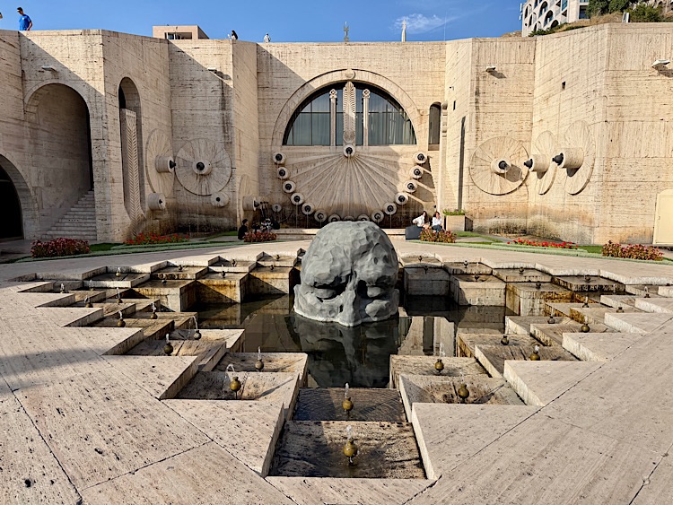 A scenic view of a modern architectural structure featuring a large stone skull sculpture in a water feature, surrounded by geometric stone pathways and a backdrop of artistic wall designs.
