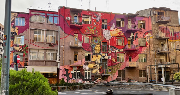 A vibrant mural on the side of a multi-story building features abstract designs, colorful flowers, and a central figure holding a traditional object, set against a pink background.