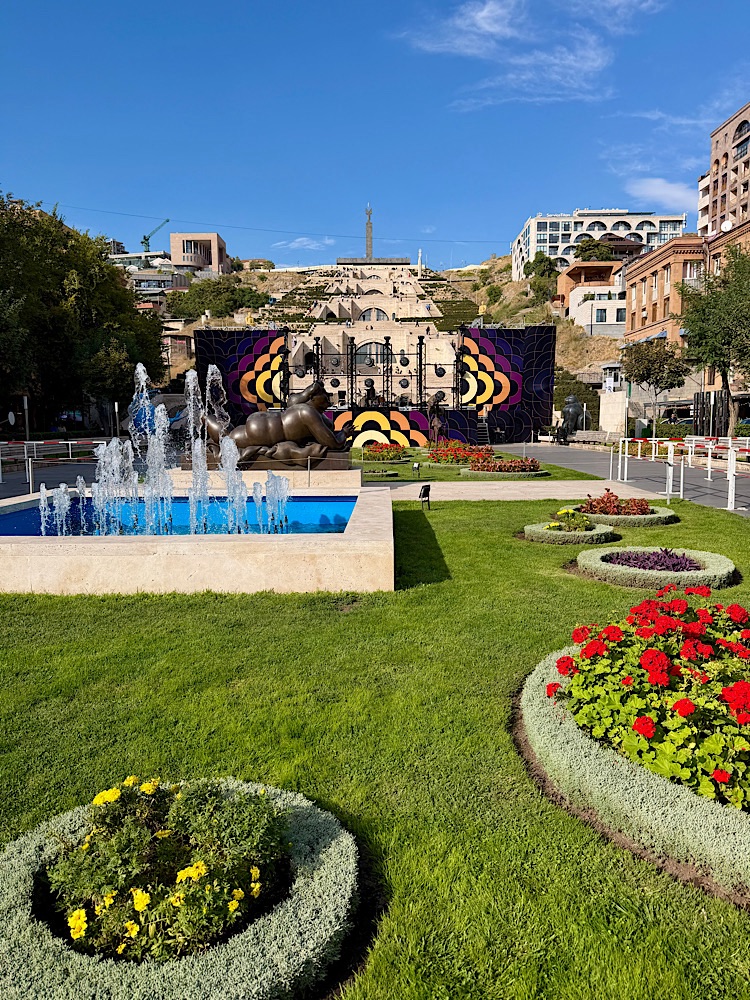 A scenic park featuring a fountain and colorful flower beds, with a backdrop of a cultural stage and hillside buildings under a clear blue sky.