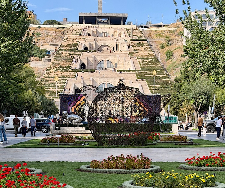 A view of the Cascade complex in Yerevan, Armenia, featuring stepped terraces with greenery and fountains, and sculptures in the foreground, surrounded by people enjoying the park.
