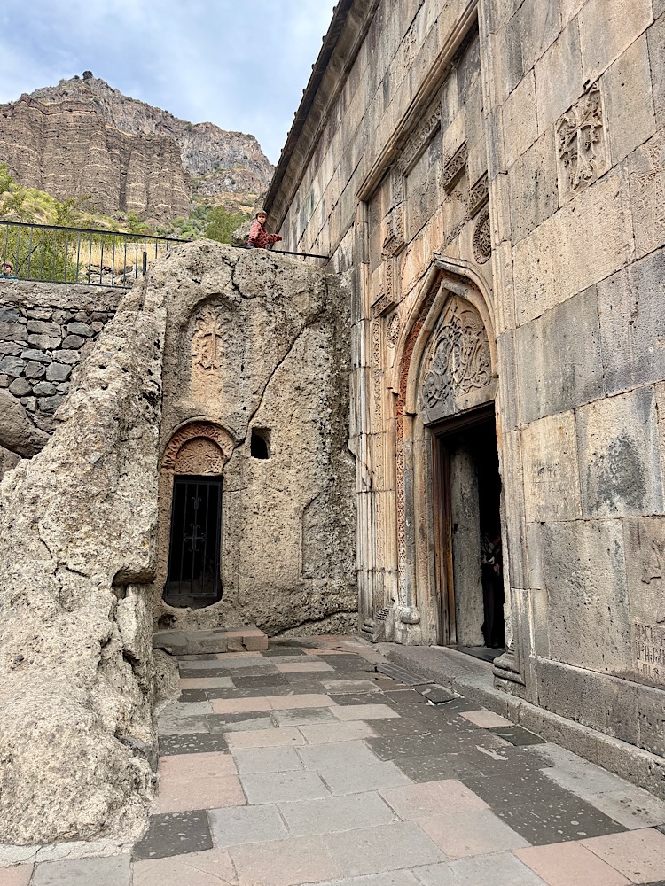 A stone structure with ornate carvings and a doorway, surrounded by rocky terrain and hills, featuring a person sitting on top of a rock ledge.
