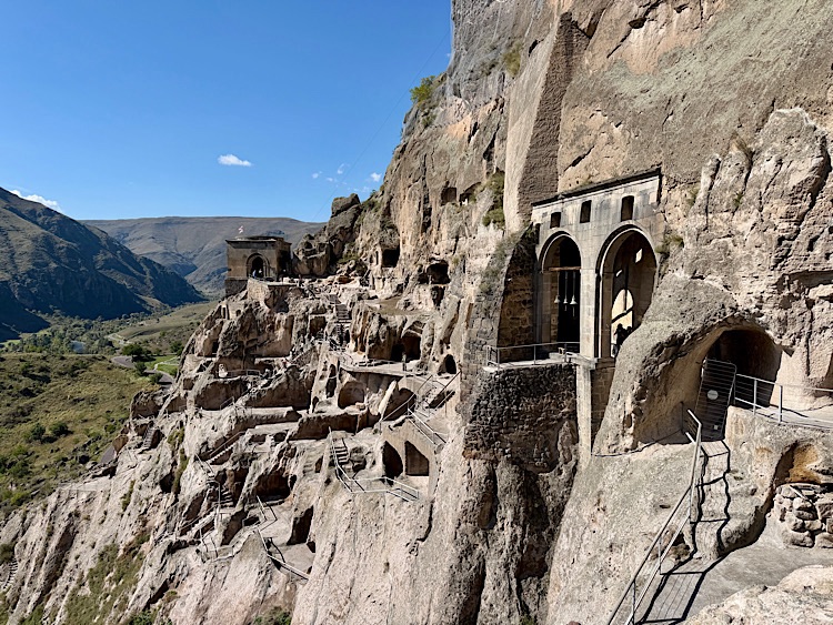Ancient rock-hewn architecture carved into a hillside, showcasing the remains of multiple caves and structures with a mountainous landscape in the background.