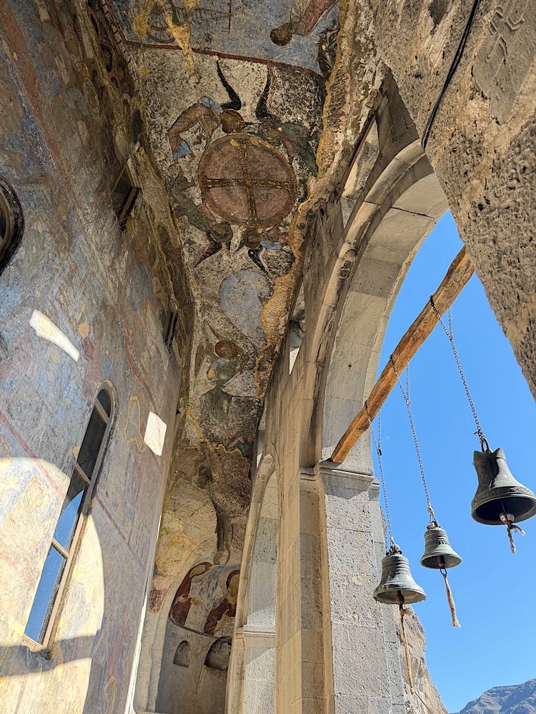 Interior view of an ancient church featuring a painted ceiling with religious frescoes, stone arches, and hanging bells.
