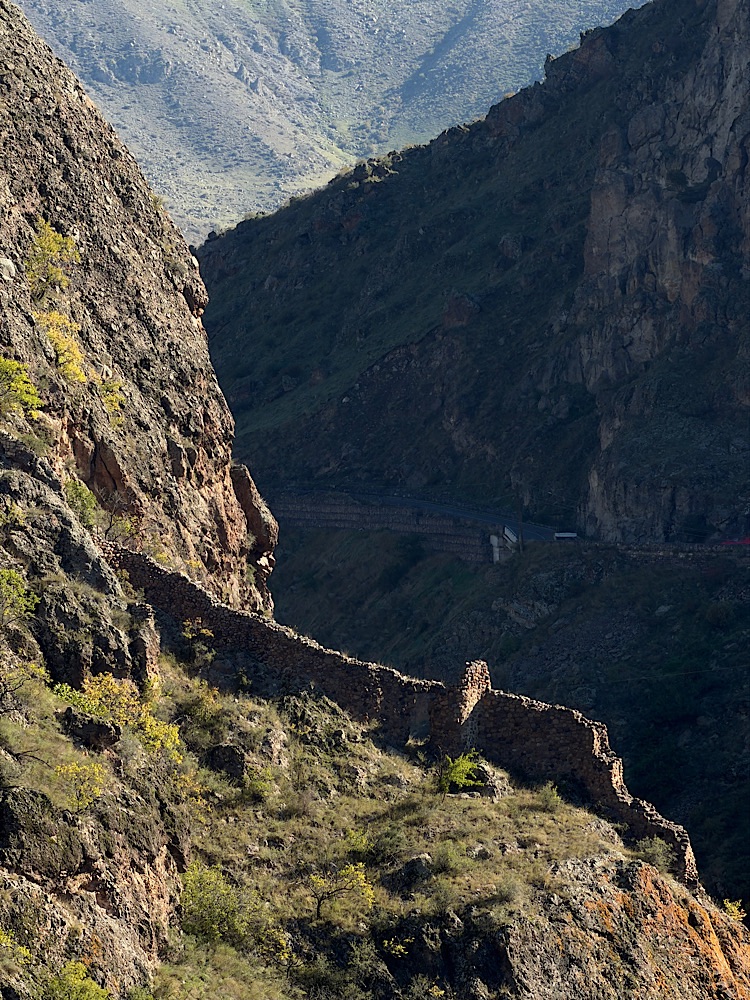 Ruins of a stone wall along a steep mountain slope with green vegetation and a winding road in the background.